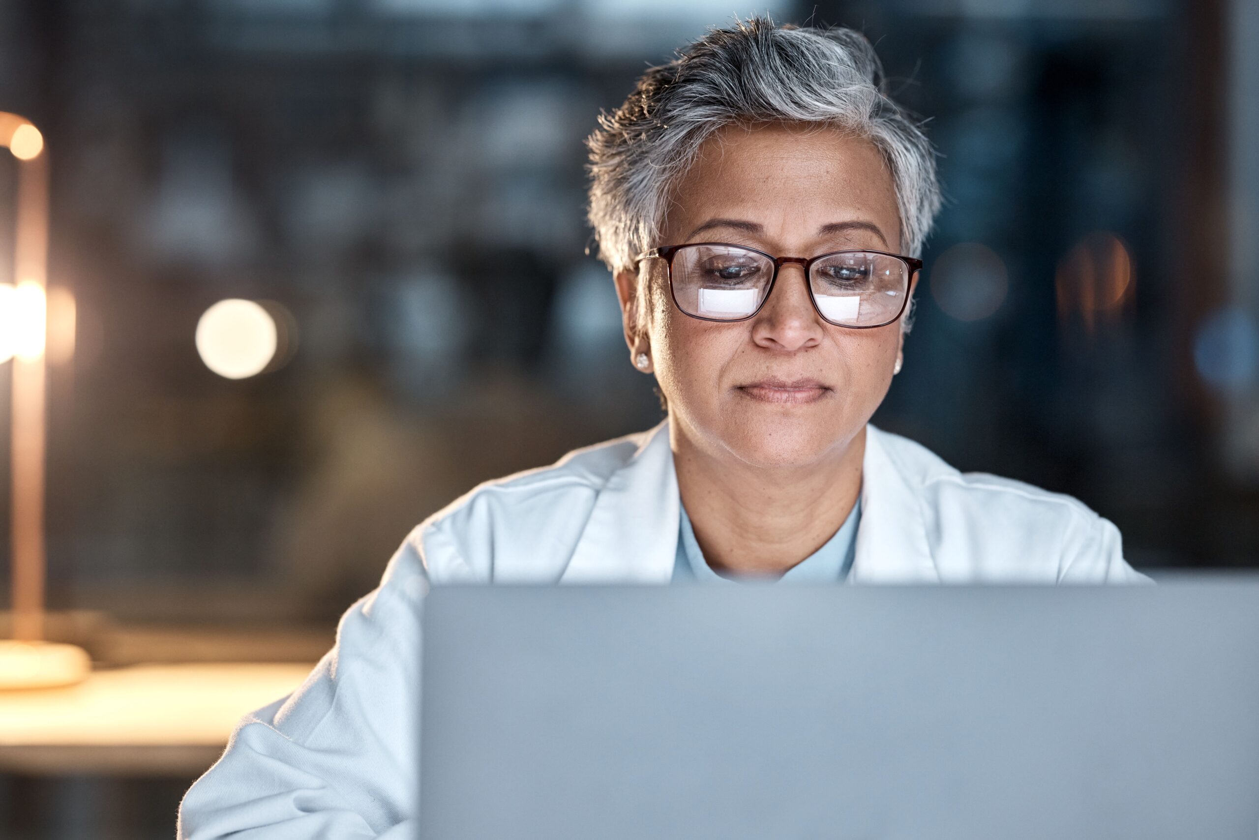 Woman,,Doctor,And,Laptop,In,Office,At,Night,At,Workplace, A doctor looking at laptop screen